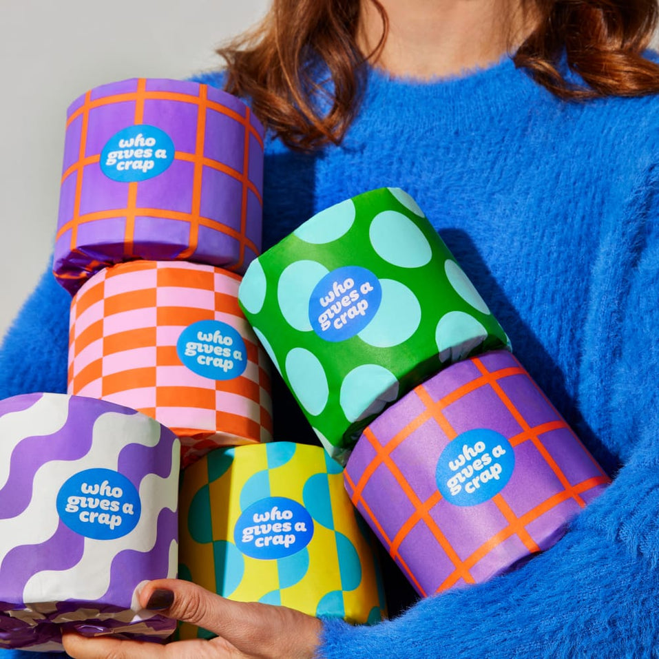 A woman in a blue sweater holds multiple wrapped colorful rolls of recycled toilet paper in her arms.