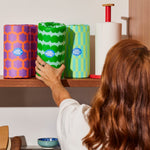 A woman reaches for a wrapped roll of kitchen towel roll that is sitting on a shelf with other wrapped and unwrapped rolls of kitchen towel.