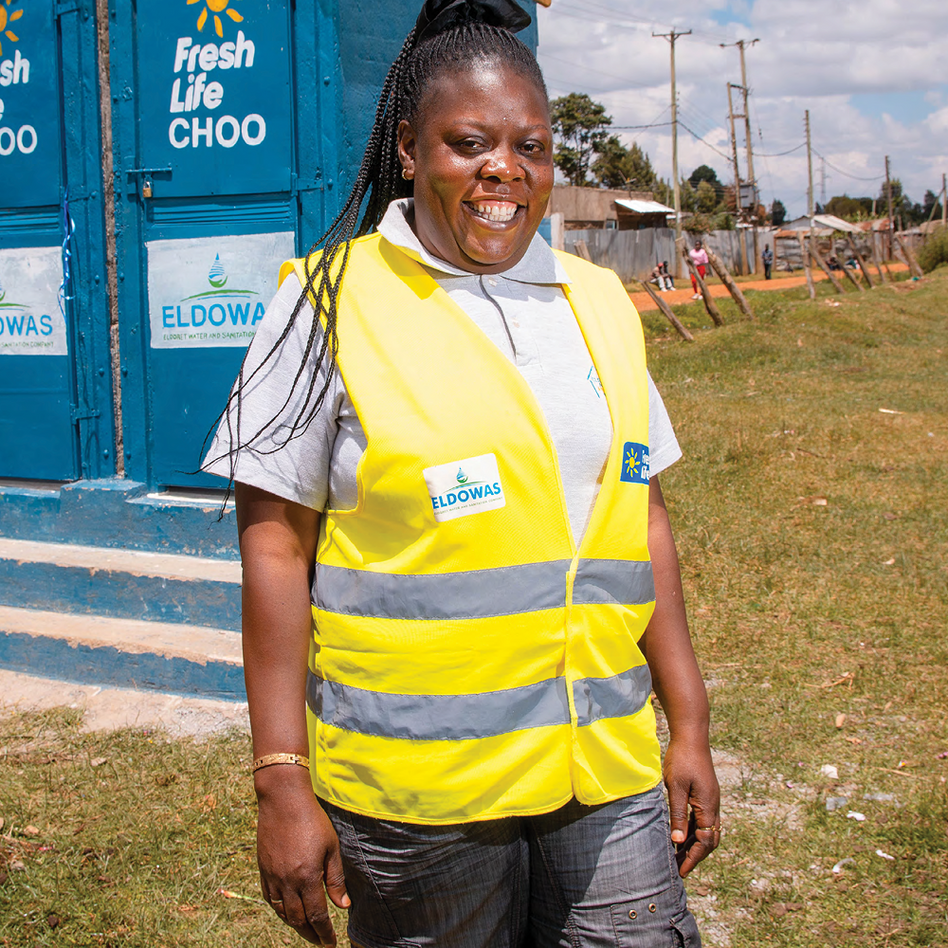 Woman in a yellow high-vis vest smiling outside a blue 'Fresh Life CHOO' toilet block in Kenya, with ELDOWAS signage on the wall